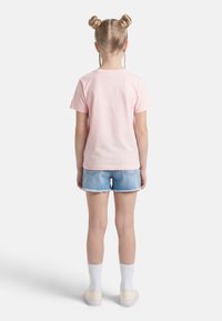 Παιδί που στέκεται με την πλάτη στην κάμερα, φοράει ανοιχτό ροζ μπλουζάκι, μπλε τζιν σορτς, λευκές κάλτσες και κρεμ αθλητικά παπούτσια, μπροστά σε λευκό φόντο.