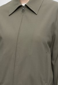 Veste vert olive avec un col pointu, une fermeture éclair frontale et une poche latérale verticale visible sur le côté droit.