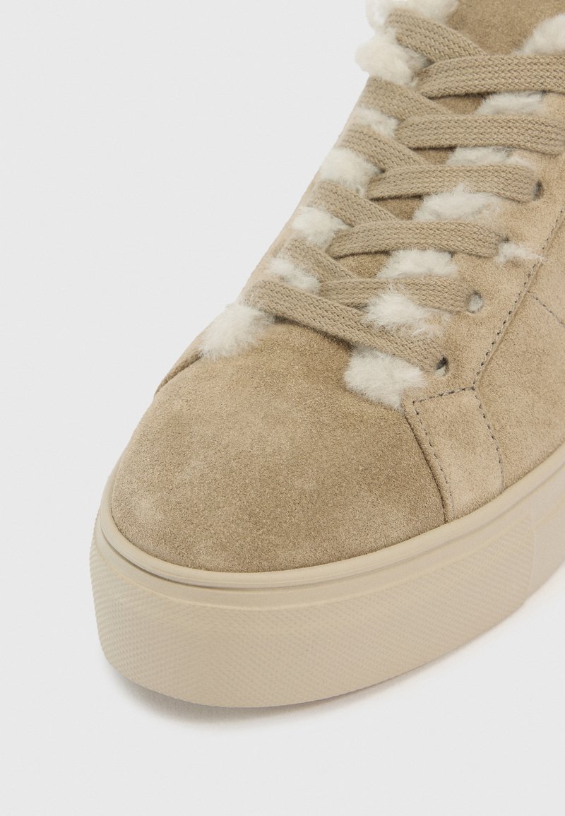 Beige Wildlederschuh mit dicker Gummisohle, beigen Schnürsenkeln und weißem, flauschigem Innenfutter, das zwischen den Ösen und rund um die Zunge sichtbar ist.