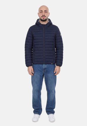 Uomo in piedi dritto, indossa una giacca trapuntata con cappuccio blu navy, jeans blu e scarpe da ginnastica bianche su uno sfondo neutro.