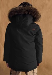 Čierna parka s kapucňou s kožušinou, s oranžovým logom, dlhými rukávmi a voľným strihom, v kombinácii s béžovými cargo nohavicami.