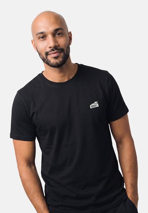Mann mit rasiertem Kopf und Bart, der ein schwarzes T-Shirt mit einem kleinen weißen "WORKS"-Logo auf der Brust trägt, steht vor einem schlichten Hintergrund.