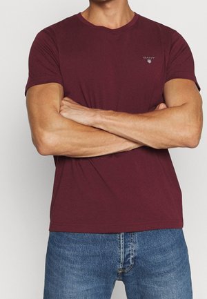 Mand iført en ensfarvet bordeaux kortærmet t-shirt og blå jeans med armene krydset over brystet.