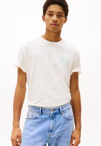 Hvid bomulds t-shirt med opkrøllede ærmer, prydet med et mintgrønt præg-logo på brystet. Brugt sammen med lyseblå denim jeans.