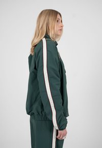 Femme aux longs cheveux blonds portant un survêtement vert foncé avec des bandes blanches et rouges, debout de profil sur un fond uni.