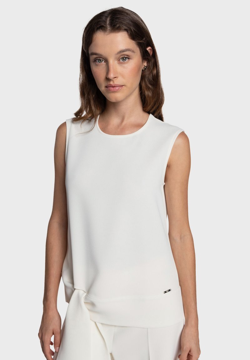 Haut blanc sans manches, tissu lisse, encolure arrondie, détail noué à la taille, accent logo subtil près de l'ourlet, design minimaliste.