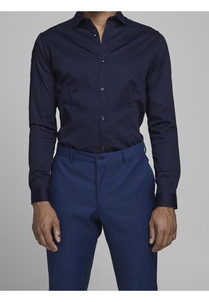 Homme portant une chemise ajustée à boutons bleu marine rentrée dans un pantalon de costume bleu assorti, sur un fond gris uni.