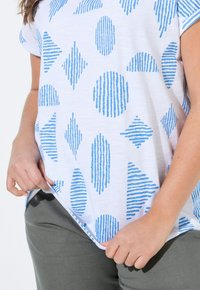 Camisa blanca de manga corta con patrones de rayas azules de varias formas, combinada con pantalones grises. La tela parece ligera y texturizada.