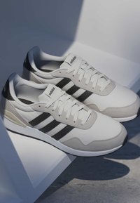 Baskets Adidas blanches et grises avec des rayures noires, des lacets blancs et un logo Adidas sur les languettes et les talons, placées sur une surface angulaire blanche.