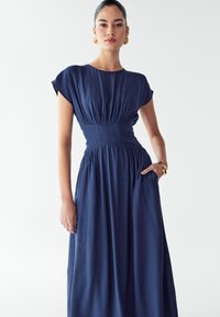 Kraljevsko modra maxi haljina s kratkim cap rukavima i strukom koji se lepo uklapa. Tkanina je glatka, sa naborima na struku i džepovima sa strane.