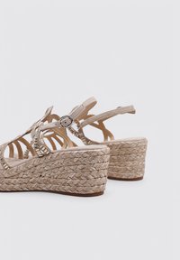 Sandali beige con zeppa e suola in juta intrecciata, dotati di fasce in pelle con ritagli e un dettaglio in metallo a forma di fibbia rotonda.