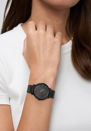 Zwart analoog horloge met een ronde wijzerplaat, een mesh bandje en minimale uurmarkeringen, gedragen om een pols tegen een witte overhemdachtergrond.