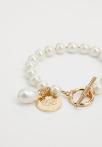 Brazalete de perlas con herrajes en tono dorado, que presenta perlas blancas redondas y una perla de forma irregular. Incluye un charm circular con iniciales grabadas.