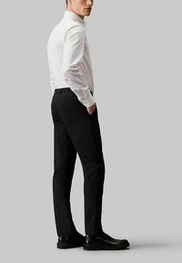 Camicia bianca a maniche lunghe abbinata a pantaloni neri su misura e scarpe nere. L'outfit presenta un design minimalista e una silhouette aderente.