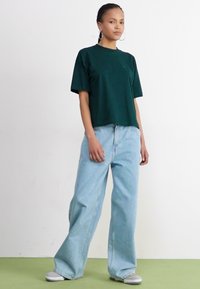 Mørkegrønn T-skjorte med korte ermer, parret med lys blå vide jeans som har en avslappet passform og minimalistisk design. Lys grå sko.