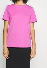 Růžové bavlněné tričko s krátkým rukávem, kulatým výstřihem a hladkou texturou. Nošeno s černou sukní, která má jednoduchý design a střih.