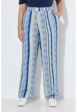 Personne portant un pantalon large à rayures verticales bleues, blanches, jaunes et vertes, ainsi que des baskets blanches, debout avec une main dans la poche.