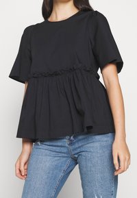 Schwarzes Peplum-Top aus Baumwollmischung mit kurzen Ärmeln, gerafftem Taillenbereich mit Rüschendetail und rundem Ausschnitt. Kombiniert mit blauen Jeans.