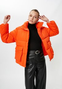 Lys orange pufferjakke med høj krave, forlommer og trykknaplukninger, båret over en sort ribbet turtleneck og sorte læderbukser.