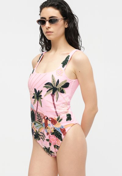 Femme portant un maillot de bain rose orné de broderies d'oiseaux tropicaux, de feuilles et de fleurs, accompagné de lunettes de soleil étroites à monture œil de chat, debout devant un fond blanc.