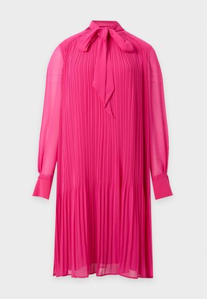 Mannequin portant une robe plissée rose vif avec un col noué, des boucles d'oreilles créoles et des bottines noires, debout devant un fond blanc uni.