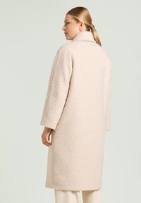 Abrigo de color beige claro, con textura, cuello alto y mangas largas. El diseño presenta un corte holgado y llega por debajo de la rodilla.