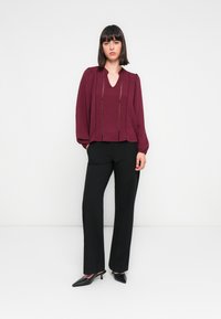 Blouse bourgogne en tissu texturé et à rayures verticales, associée à un pantalon noir et des escarpins noirs pointus. Coupe décontractée avec des manches ballon.