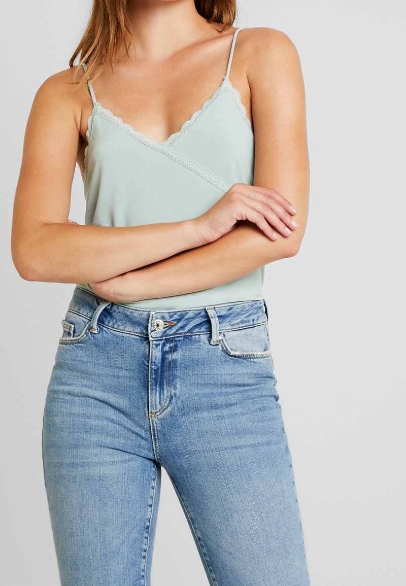 Femme portant un caraco vert clair bordé de dentelle rentré dans un jean bleu clair taille haute, bras croisés sur la poitrine, sur un fond uni.