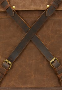 Sac en toile marron avec des sangles en cuir noir entrecroisées, fixées par des boucles en laiton, mettant en valeur une texture robuste et un design solide.