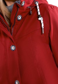 Rød vandtæt jakke med en glat overflade, sølv lynlås, justerbar snørelukke hætte og knapdetaljer med et logo.