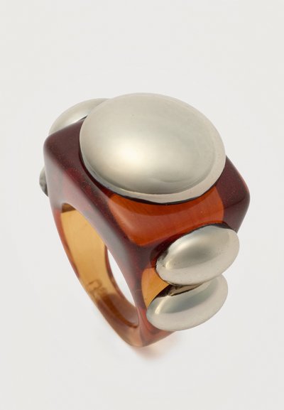 Τετράγωνο δαχτυλίδι από κεχριμπαρένια ρητίνη με μεγάλες, λείας επιφάνειας ασημένιες κυκλικές και οβάλ μεταλλικές διακοσμήσεις σε κάθε πλευρά.