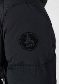 Zwarte pufferjack met een ronde, geprofileerde patch met een logo op de bovenarm. De stof lijkt soepel en geïsoleerd.