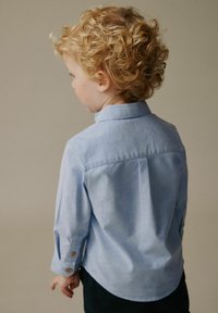 Cămașă albastru deschis cu mâneci lungi și guler cu nasturi, având un pli în partea din spate și nasturi din lemn la manșetă, purtată de un copil cu spatele la privitor.