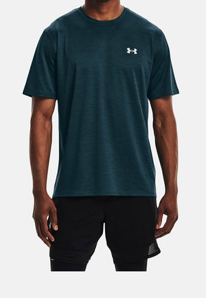 Mężczyzna w ciemnozielonym sportowym t-shircie z krótkim rękawem z białym logo na klatce piersiowej i czarnych szortach, stojący na jasnym, jednolitym tle.