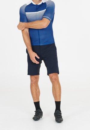 Hombre con maillot de ciclismo de manga corta azul y blanco, pantalones cortos de ciclismo negros, calcetines negros y zapatillas de ciclismo negras sobre fondo blanco.
