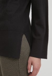 Maglione scuro con lavorazione a maglia texturizzata, dotato di aperture laterali e maniche lunghe, abbinato a pantaloni a scacchi in marrone e beige.