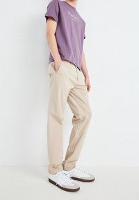 T-shirt in cotone viola con logo bianco, abbinata a pantaloni beige chiari e sneakers bianche con dettagli grigi. Outfit semplice e casual.