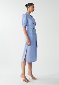 Robe bleu clair, rayée, mi-longue avec des manches ballons, un design enroulé, des boutons, des poches et des fentes latérales, associée à des sandales blanches.