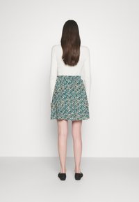 Un haut à manches longues blanc associé à une jupe turquoise à motifs ; taille élastique, longueur genoux, et des chaussures plates noires. Texture lisse sur l'ensemble.