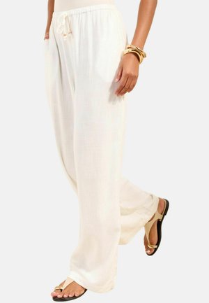 Nő laza, fehér, húzózsinóros nadrágban, bézs szandálban és aranykarkötőkben, egyik lábát kissé felemelve áll.