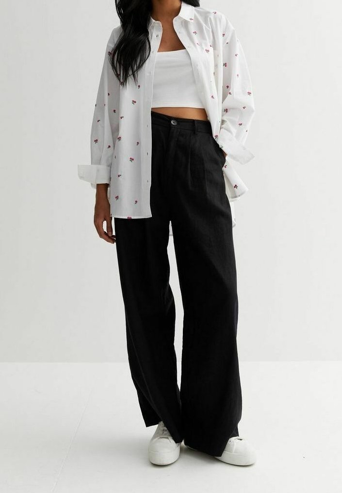 Personne portant une chemise blanche à fleurs boutonnée sur un crop top blanc, un pantalon noir à jambes larges et des baskets blanches, debout devant un fond uni.