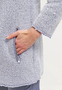 Lätt, stickad jacka i ljusblått och vitt, med en dragkedjeficka och fransade kanter på ärmsluten och nederkanten. Strukturerad tyg.