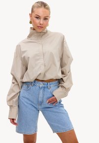 Femme aux cheveux tirés en arrière portant de grandes boucles d'oreilles dorées, un chemisier beige à col haut et un short en jean bleu clair, posant avec une main sur la hanche.