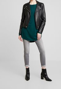 Schwarze Leder-Motorradjacke über einem grünen T-Shirt, kombiniert mit grauen Jeans und schwarzen Stiefeletten. Die Jacke ist mit silbernen Reißverschlüssen und Druckknöpfen versehen.