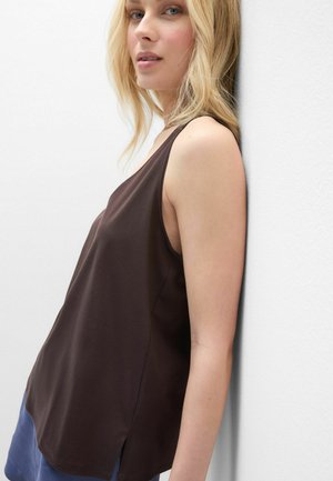 Mladá žena s blond vlasy se opírá o bílou zeď, má na sobě tmavě hnědý top bez rukávů a modré kalhoty, a dívá se mírně stranou.