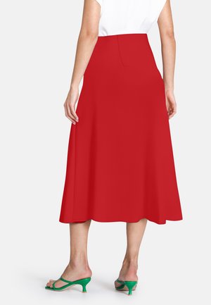 Frau mit rotem Midirock, weißem Oberteil und grünen Sandalen mit niedrigen Absätzen, die mit dem Rücken zur Kamera steht.