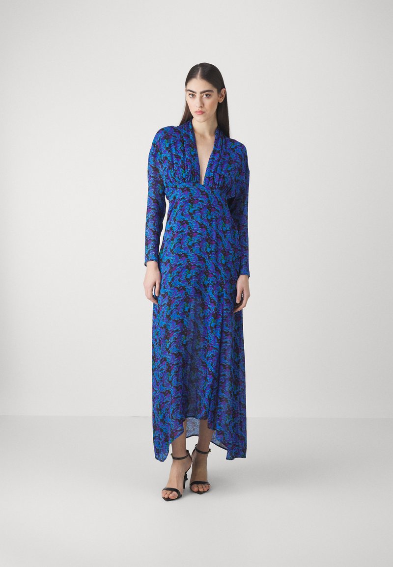 Robe longue bleue avec un motif floral comportant des détails violets et verts. Décolleté en V et manches longues. Portée avec des sandales noires à lanières.