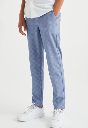Homme portant un pantalon à carreaux bleu clair, des baskets blanches et une chemise blanche boutonnée, debout les mains dans les poches.