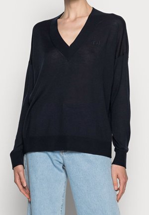 Kvinde iført en løs sort V-hals sweater med ribbet kant ved ærmer og bund, kombineret med lyseblå jeans, stående foran en ensfarvet baggrund.
