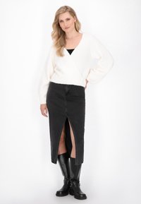 Femme blonde portant un pull enveloppant blanc, une jupe mi-longue fendue noire et des bottes noires montantes, se tenant avec une main sur la hanche sur fond blanc.